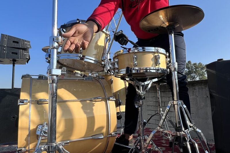 A student drummer checking cymbals on an outdoor stage rig, PA and lighting truss behind him